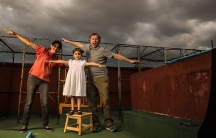 A woman, a child and man spread their arms out like airplanes on the roof of a building as they pose for a silly family photograph. Behind them are dark storm clouds.