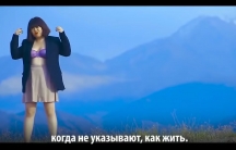 A woman wears a lacy bra and a blazer as she stands in front of a sweeping mountain vista in this screen grab from a music video. Below, a line from the song is spelled out in Kyrgyz.