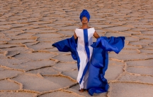 Malian singer Fatoumata Diawara is shown in a bright blue and white dress standing in the desert.