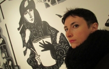 Victoria Lomasko is shown in this portrait photo with black coat standing in front of an illustrated wall.