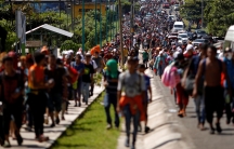 Central American migrants are shown walking on a road and sidewalk spreading the length of the highway in frame near the border with Guatemala.