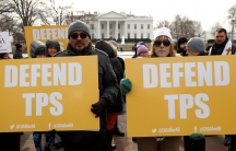 People hold bright yellow signs reading "Defend TPS" in front of the White House in Washington, DC.