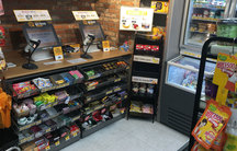 Two computers with scanners are set up on a counter in a convenience store, surrounded by packaged snacks and rows of gum and mints.