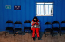 A woman sits in the middle of a row of chairs, her legs out in front of her, looking at her cell phone.