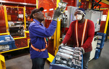 One person inspects a piece of metal, while another person holds a cart containing a more of the same pieces of metal. 