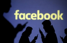 Silhouettes of people looking at their mobile phones in front of Facebook's logo. 