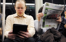 A commuter reads on a Kindle e-reader while riding the subway in Cambridge, Mass. Neuroscience says the way his brain treats reading on the Kindle is different than the way the brain processes the newspaper next to him.