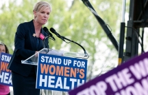 A blond woman is speaking into a microphone at a podium with a sign on the front that says 