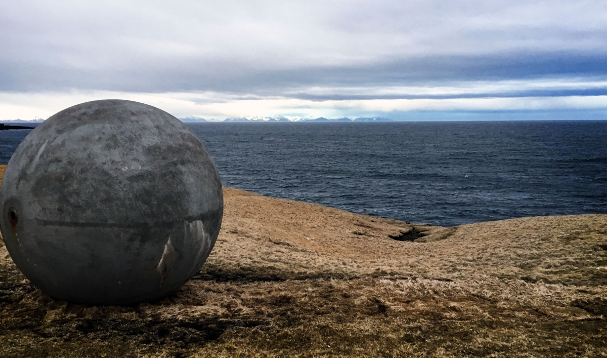 A large concrete sphere sits on a brown hill overlooking a slate blue sea.