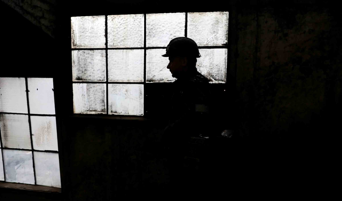 A man wearing a hard hat is silhouetted against old heavy windowpanes in a factory.