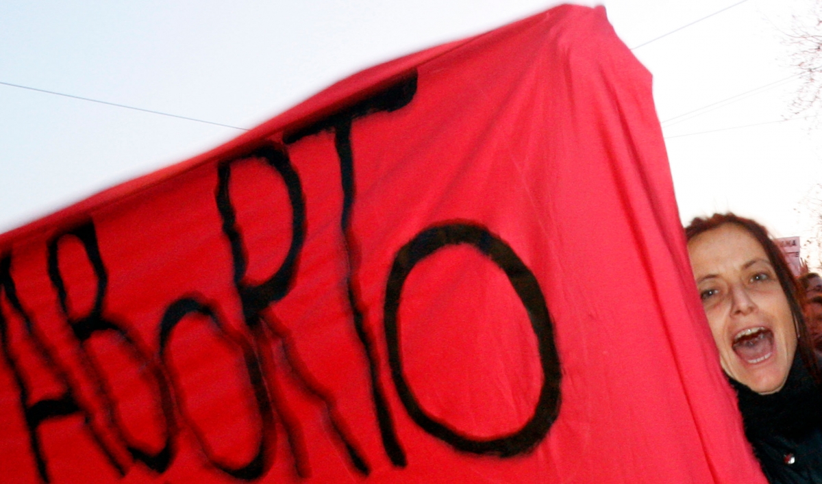 A woman's face looks out from behind a red protest banner that says "aborto"