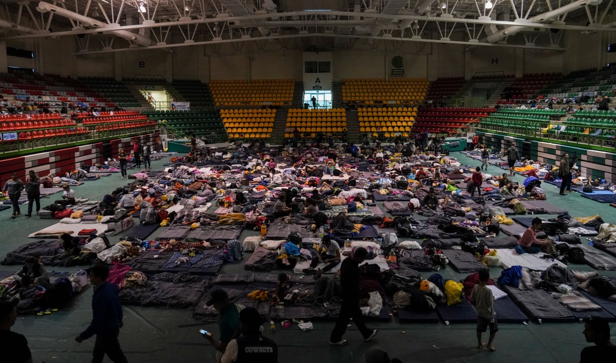 Migrants sitting and laying on mats are shown spread across a high school gymnasium floor.
