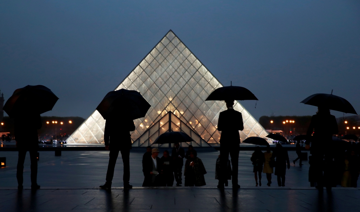 Silhouetted people with umbrellas stand in front of the Louvre pyramid at night.
