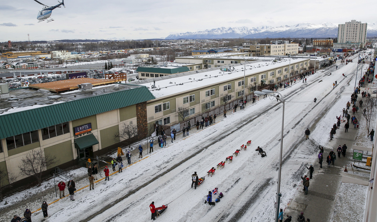 A cityscape of Anchorage, Alaska with dog-sled racers and mountains