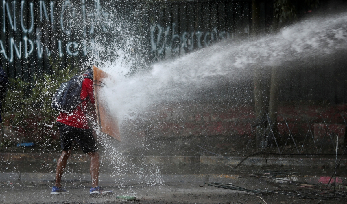 A demonstrator wearing a red shirt and shorts is shown with a makeshift shield held up protecting themselves from a police water canon.