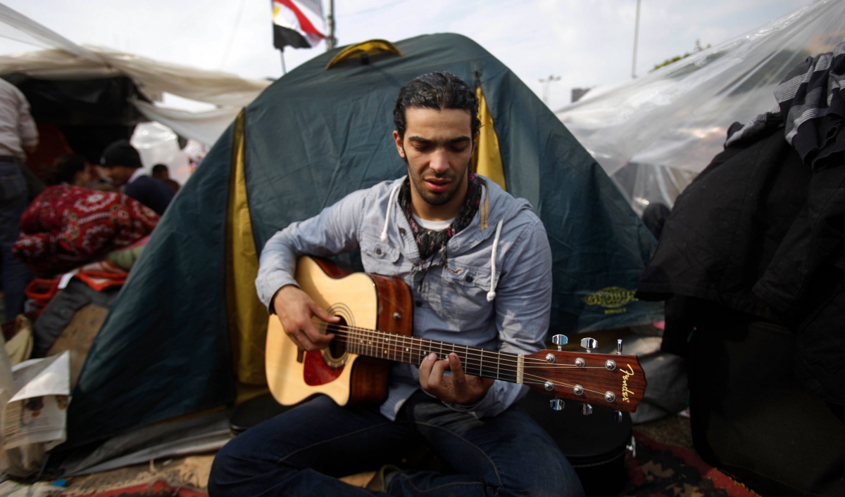 A man is sitting cross-legged in front of a tent and holding a guitar.