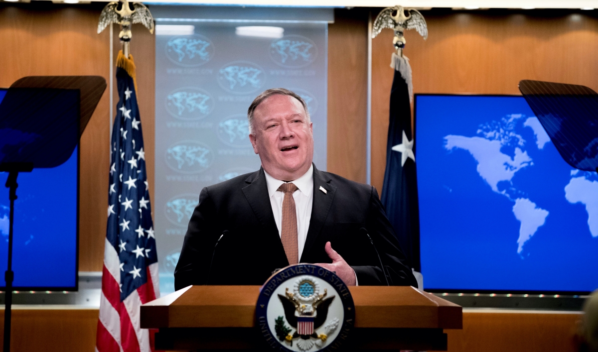 A man wearing a suit stands at a State Department podium between two flags
