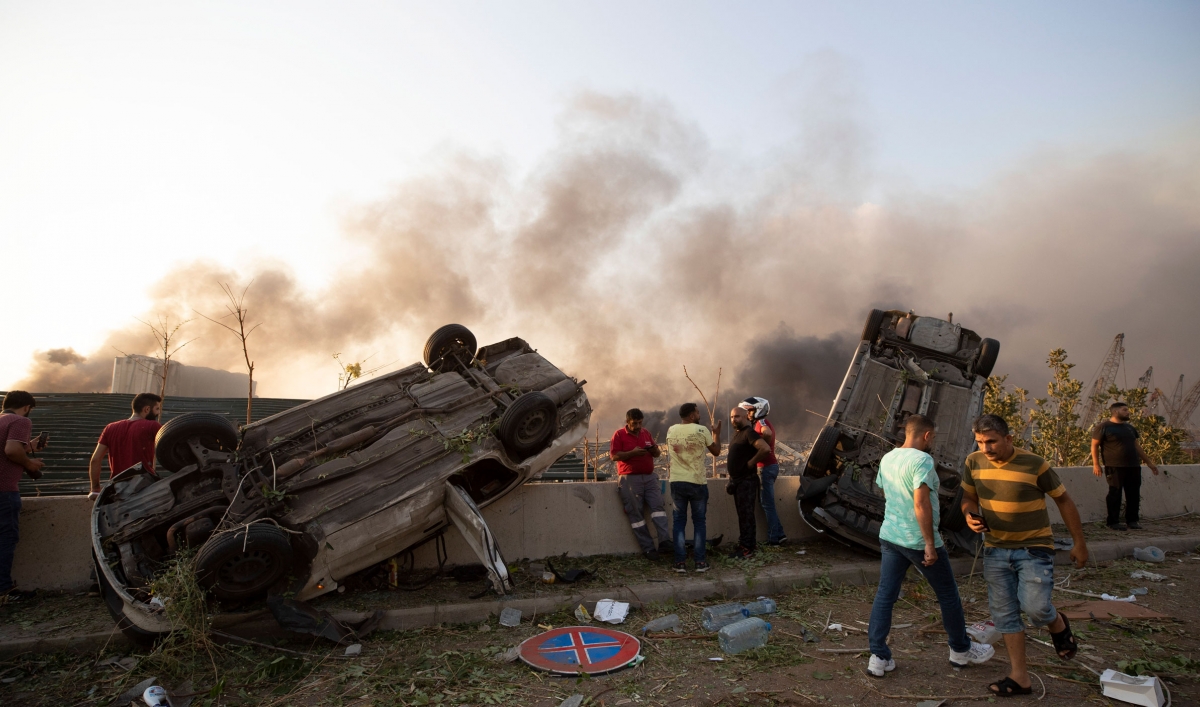 Smoke is seen rising in the air with several people standing and two cars are flipped over a barrier.