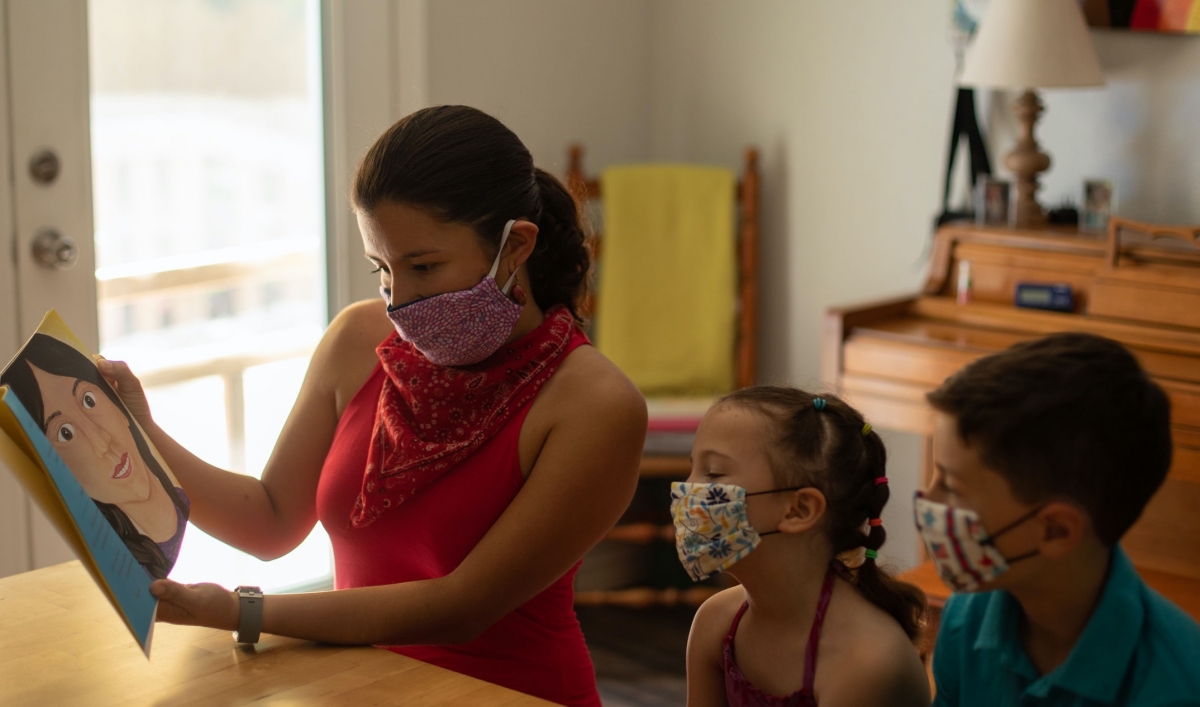 A woman wears a mask and reads a book to two children, also wearing masks.