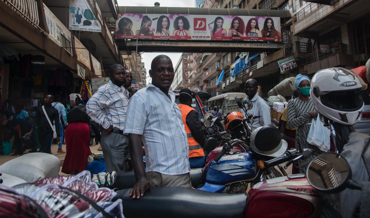 A man stands in front of many motorcyles and drivers wearing helmets outside in front of an overpass.