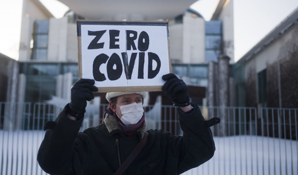 A man holds a sign that says "zero COVID" wearing a winter jacket and hat outside in front of chancellery in Berlin.