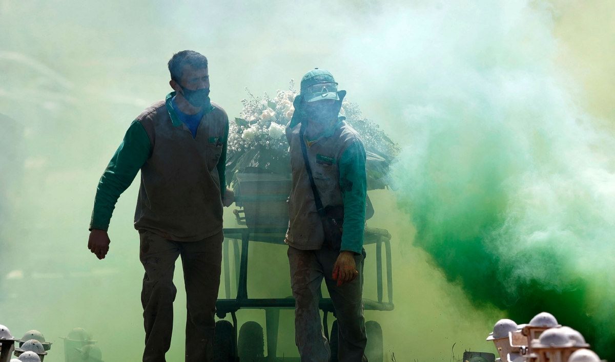 Two cemetery workers are shown pulling a casket on wheels with ceremonial green smoke surrounding them.