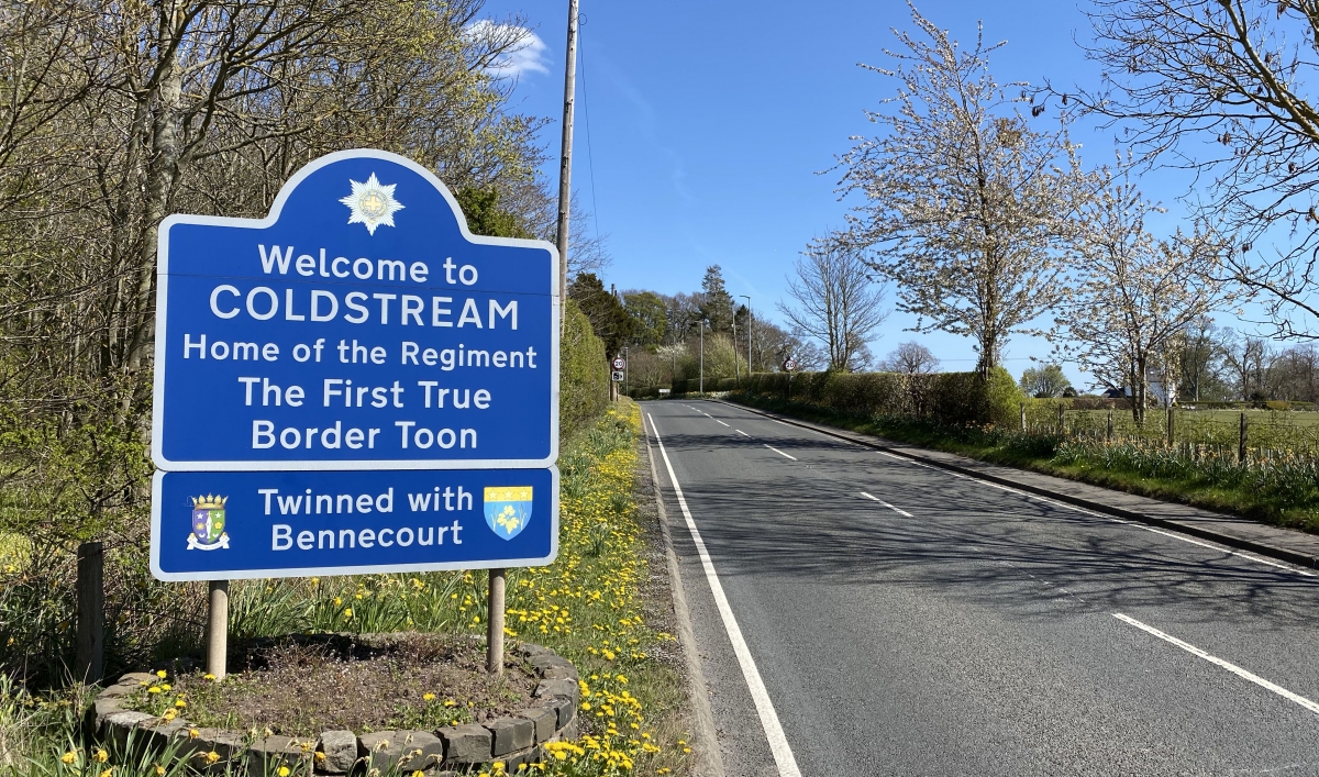 A blue border town sign along a road with foliage.