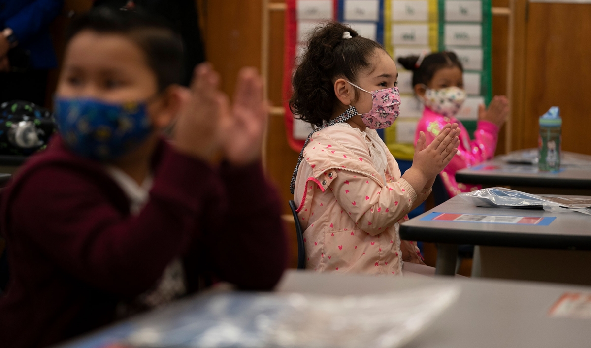 Children in pink and maroon jackets applaud in their classroom