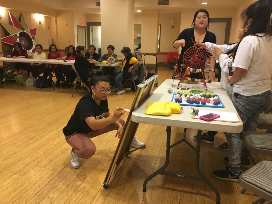 Elderly people sit at a long table, as younger people set up a game of bingo