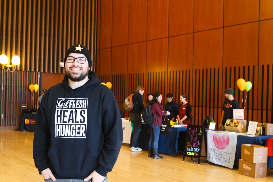 Man standing in auditorium in front of tables, in CalFresh sweatshirt