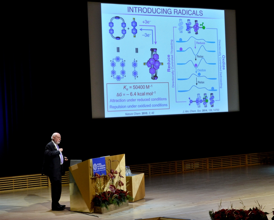Man at podium on stage pointing to powerpoint slide with scientific information