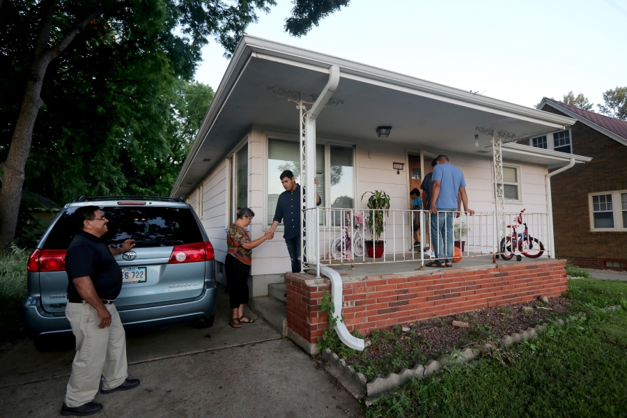 Van on left in driveway of home, man walking toward front porch, young man helping woman up stairs to porch, other entering home