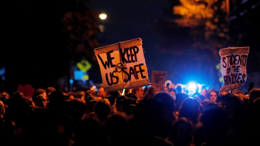 A crowd of people is illuminated by the blue of a police light and a sign says "We keep us safe" and with a symbol for no cops.