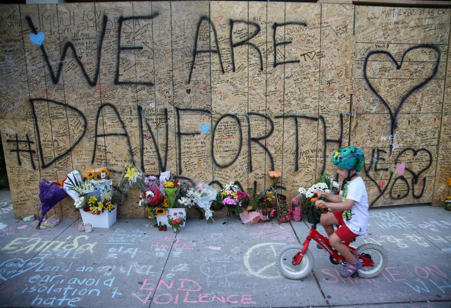 A small boy rides a bicycle past a plywood wall that says "We are Danforth" inscribed with messages, hearts and flowers.