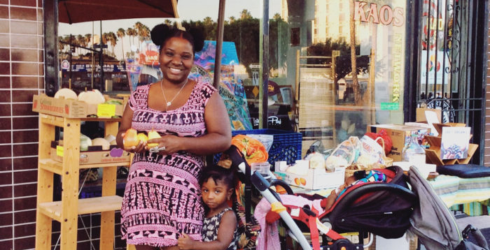 Shoppers at a recent SÜPRMARKT pop-up. A young woman in a pink dress holds veggies in her hand with a child standing by her side.
