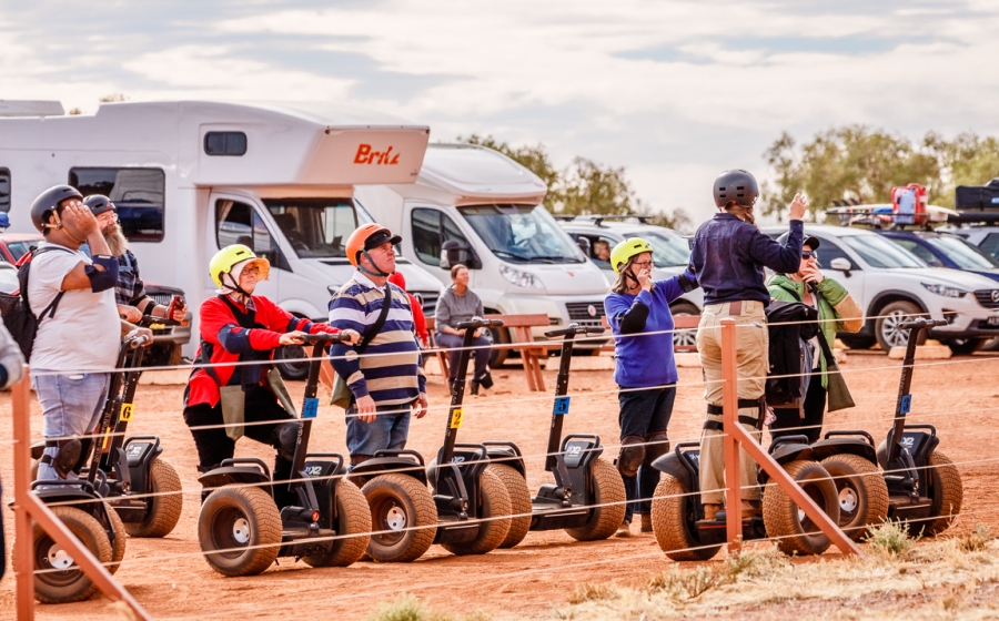 Don't want to walk around the base of Uluru? How about a tour on a Segway? With the rock closing to climbers in 2019, Parks Australia and local tourism operators are developing alternative activities. Tourists stand on segues wearing helmets looking off at Uluru as the sunset brings vast color changes and shadows to the face of the rock.