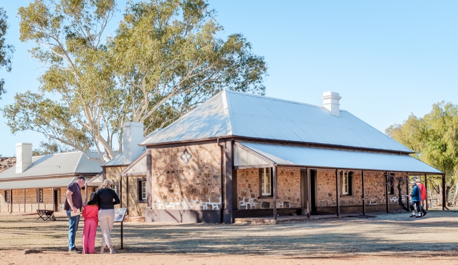 Telegraph Station is an historic building from early colonial times just outside of Alice Springs. It was one of 12 stations along the Overland Telegraph Line, and was closed in 1932. Telegraph Station an historic building is shown with a small family looking at a sign in the foreground.