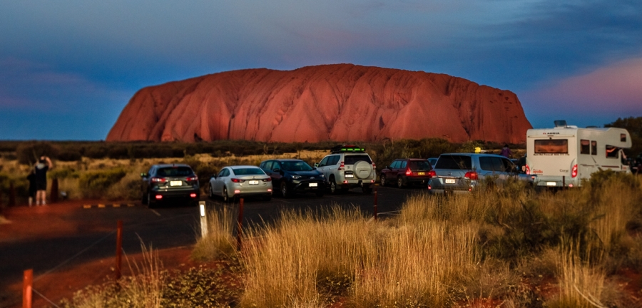 Tourists gather at this evening viewing spot to see Uluru as the sunset brings vast color changes and shadows to the face of the rock. Cars are shown in the foreground near the base of Uluru, cast in red light from the sunset.