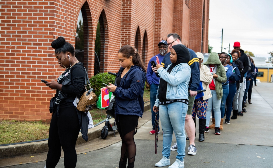 A line of people wraps around a brick building. Most people stand in line, looking at cell phones.