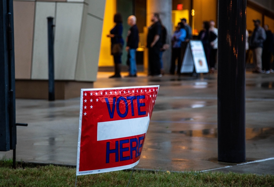 A red sign says "Vote here." In the background, a line of voters extends out the glass doors of a building.