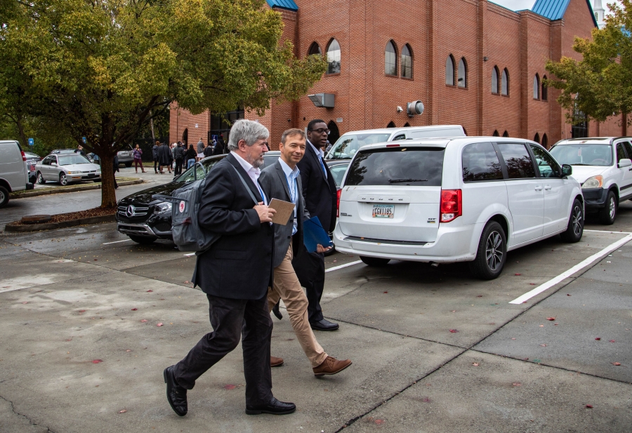 Three men walk through a parking lot. Behind them is a line of people waiting to vote at a church.