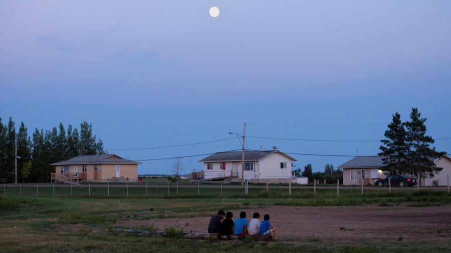 Children from the Cote First Nation sit at the campground set up by the First Nations Indigenous Warriors and the American Indian Movement on the Cote First Nation, near the town of Kamsack, Saskatchewan, Canada, Aug. 6, 2017. Children sit on a log under the moonlight in a field surrounded by buildings.