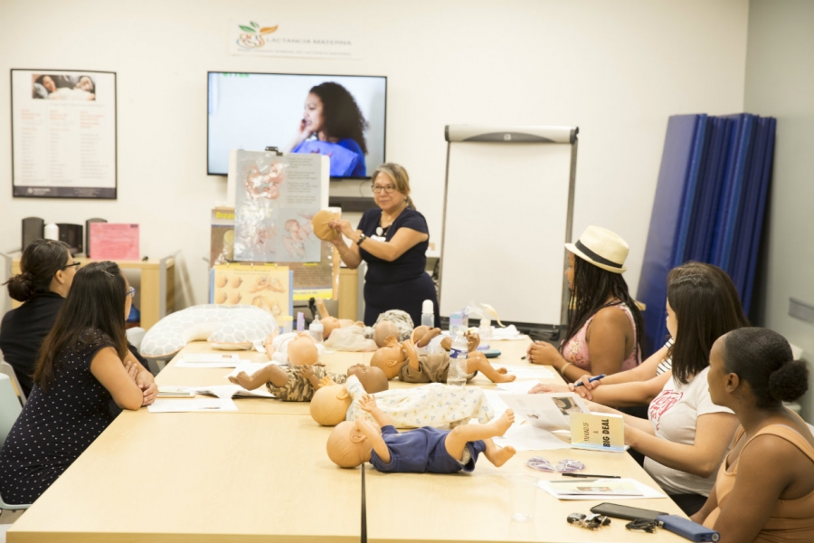 A group of women sits around a table listening to a speaker. Dolls are on a table.