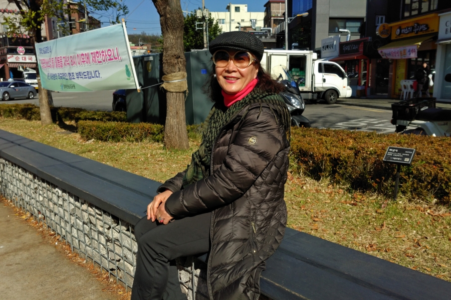 Lee Soon-ja, 54, from Busan, South Korea. Woman in black coat and hat poses for camera on street ledge.