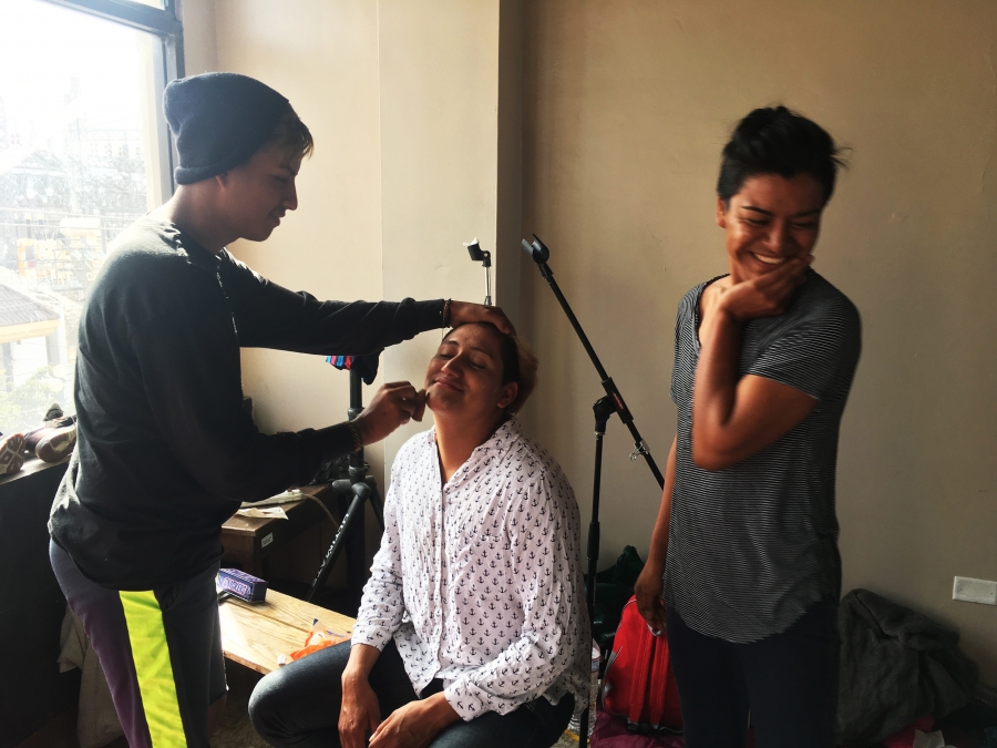 A few migrants prepare for the weddings by putting on makeup. One sits in a chair while another applies makeup and another giggles in the corner