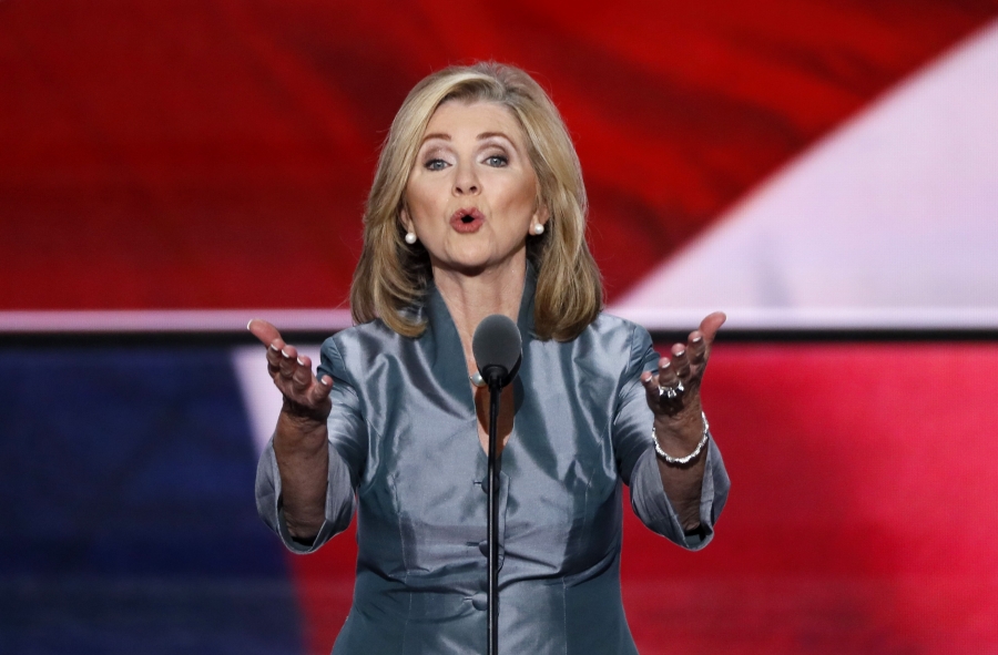 speaks behind a podium with her arms outstretched toward the audience