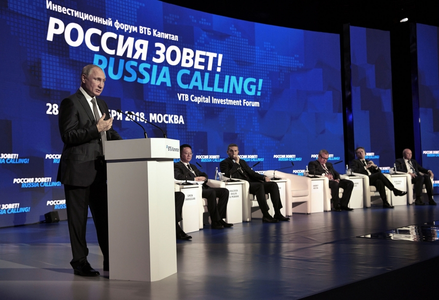 Russian President Vladimir Putin stands at a podium. To his right are other officials seated on a stage.