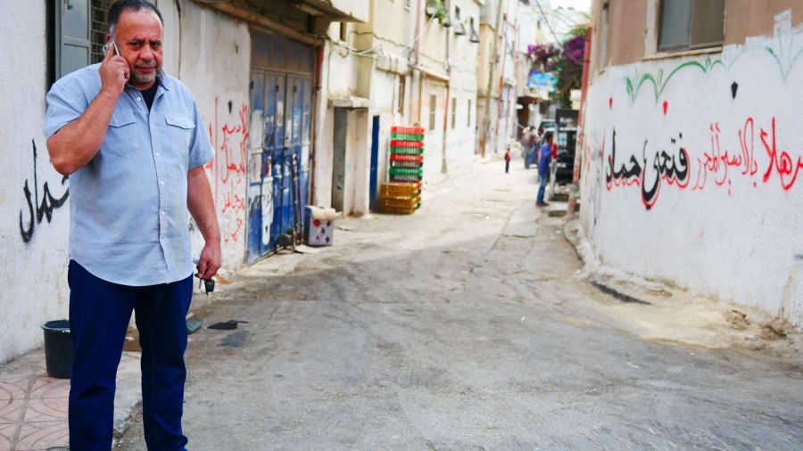 Taha al-Biss, a leader in Am'ari camp, walks along a street riddled with cracks and frequently inundated with sewage. A man wearing a blue shirt and jeans walks on the street with his phone near his ear.