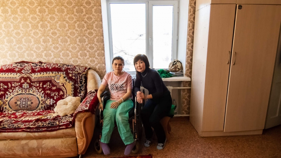 Aigul Suleimenova sits with her daughter, Assee, who has a condition that weakens her nervous system. “She was born like that,” Aigul said. Assee, now 29, spends her days in a wheelchair and is under constant supervision. A young woman sits in a wheelchair next to her mom in the living room.