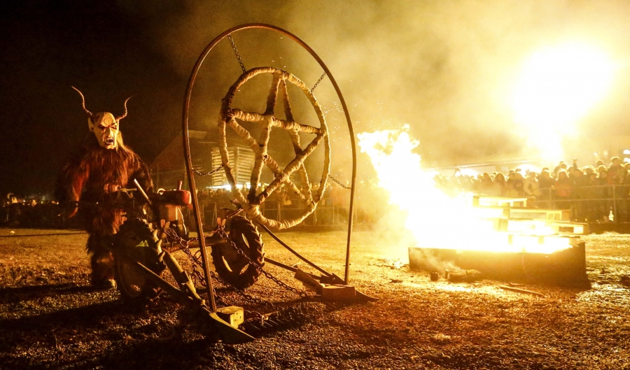 A man dressed in a traditional Perchten costume and mask performs in a field next to a bon fire